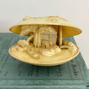 May include: A cream-coloured clam shell ornament. The interior is carved with a miniature village scene, including a house, trees, a small animal, and a bridge. The shell is open, displaying the intricate details of the miniature landscape.