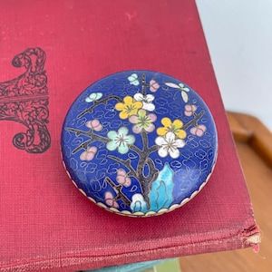 May include: A round, blue enamel trinket box with a floral design. The box features a dark blue background with a tree and colorful flowers in yellow, pink, and white. The box is resting on a red surface.