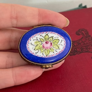 May include: An oval, blue trinket box with a floral design. The lid features a central oval with a pink rose and green leaves on a white background, surrounded by a blue border. The box has a gold-colored metal frame.