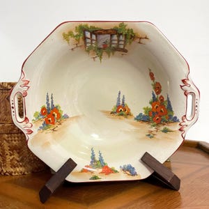 May include: Cream-colored, octagonal serving dish with a red rim and handles. The dish features a painted landscape with a window, flowers, and foliage. The dish is supported by two wooden stands.