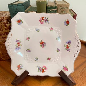 May include: A square, white porcelain serving plate with a scalloped edge and floral designs. The plate features a raised central square and is decorated with clusters of red, pink, purple, and blue flowers. Gold trim accents the edge.
