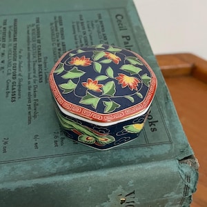 May include: A small, hexagonal trinket box with a navy blue background and floral design. The lid features red and yellow flowers with green leaves. The box has a red and white border and is likely made of ceramic.