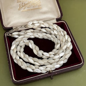 May include: A white mother of pearl necklace with a silver clasp in a brown velvet lined box. The necklace is made of oval shaped beads.
