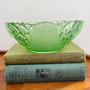 May include: A green glass bowl with a frosted finish and a leaf design. The bowl is sitting on top of two books.