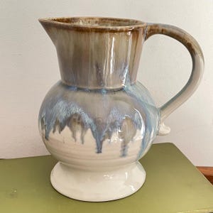 May include: A white ceramic jug with a brown glaze and blue accents. The jug has a round body and a wide, flared spout. The handle is attached to the side of the jug and curves up and over the top.