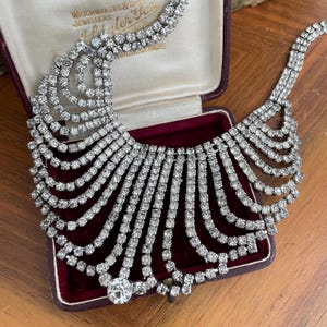 May include: A vintage rhinestone necklace with a striking, cascading design. The necklace is made of multiple strands of sparkling clear rhinestones. It is presented in a burgundy jewellery box, showcasing its elegant, eye-catching effect.