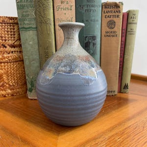 May include: A handcrafted ceramic vase with a textured, ribbed design. The vase features a muted colour palette of blue, grey, and brown, with a flared neck. The vase is displayed on a wooden surface, with vintage books in the background.