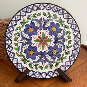 May include: A round, hand-painted ceramic plate with a black rim. The plate features a floral design with blue flowers, green leaves, and a red and pink center. The plate is displayed on a wooden stand.