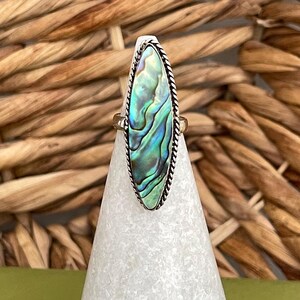 May include: A silver ring with an oval abalone shell displaying iridescent blues, greens, and purples. The shell is framed by a twisted silver rope design. The ring is displayed on a white cone against a woven background.