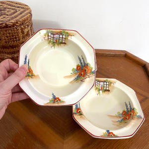 May include: Two octagonal cream-colored bowls with a red trim. Each bowl features a painted scene of a window, red poppies, and blue flowers. The bowls are displayed on a wooden surface.