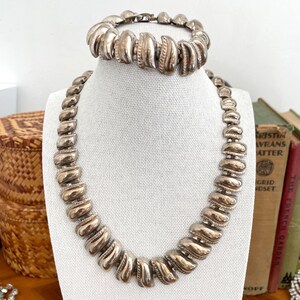 May include: A silver-coloured necklace and bracelet set. The necklace and bracelet feature a series of curved, ridged, and textured links. The jewellery is displayed on a white mannequin against a backdrop of books and a woven basket.