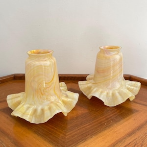 May include: Two vintage glass lampshades with a ruffled edge. The shades have a yellow and cream swirl pattern. The lampshades are sitting on a wooden surface.