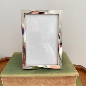 May include: A rectangular picture frame with a polished silver-tone metallic finish. The frame features a distinct twisted or warped design along the vertical sides, creating a reflective surface that catches light. The center contains a white insert.