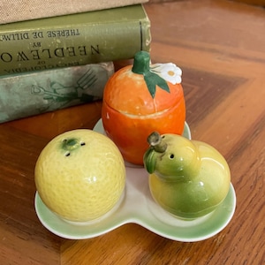 May include: A ceramic fruit-shaped salt and pepper set. The set includes a yellow lemon, a green pear, and an orange with a white flower on top. The pieces sit on a light green tray. Vintage kitchen decor.