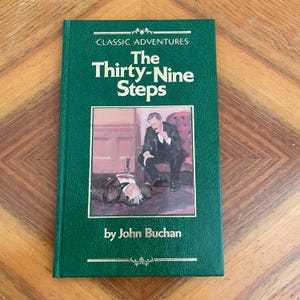 May include: A green hardcover book, "The Thirty-Nine Steps" by John Buchan, with gold lettering. The cover features a colour illustration of two men. Part of the "Classic Adventures" series.