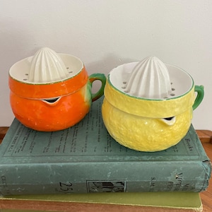 May include: Two ceramic citrus juicers, one orange and one yellow, resembling an orange and a lemon. Each has a white reamer and a green handle. The juicers are sitting on top of a stack of books.