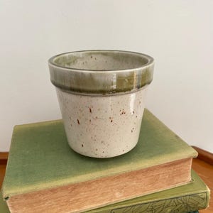 May include: A cylindrical ceramic planter with a speckled cream-coloured body and a smooth, glossy olive-green glazed rim. The vessel features a straight-sided design with a wide opening, showcasing a natural, earthy aesthetic with visible brown speckles.