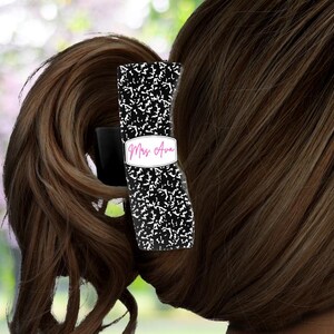 May include: A black and white patterned hair clip with the text "Miss Ava" in pink script. The clip is holding a brown ponytail. The background is a blurred green and pink.