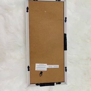 May include: White wood picture frame with black metal accents. The frame has a brown cardboard backing and a hanging hook.