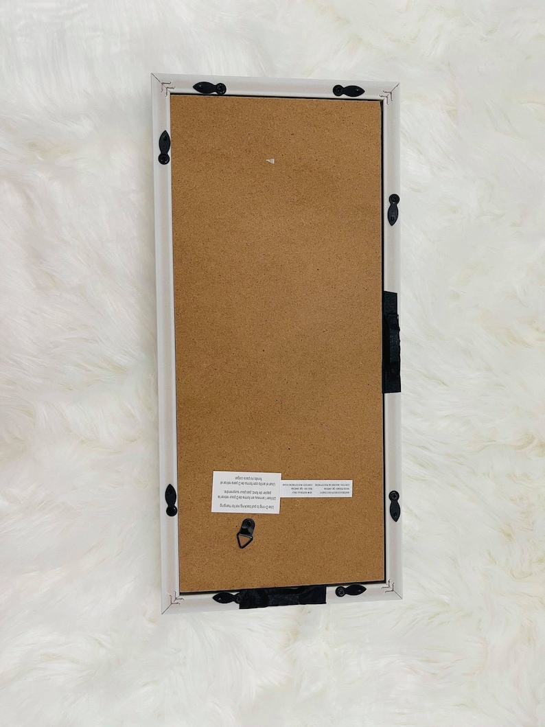 May include: White wood picture frame with black metal accents. The frame has a brown cardboard backing and a hanging hook.
