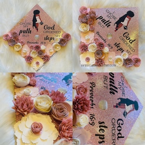 Op de afbeelding: Een afstudeerhoed versierd met roze en witte papieren bloemen. De hoed heeft een glinsterende roze achtergrond en de tekst "Ik koos mijn pad, maar God heeft mijn stappen geordend Spreuken 16:9".