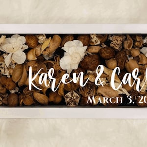 May include: A rectangular white framed shadow box filled with dried botanicals in shades of brown, beige, and white. The names "Karen & Carlo" and the date "March 3, 2021" are written in white script across the center.
