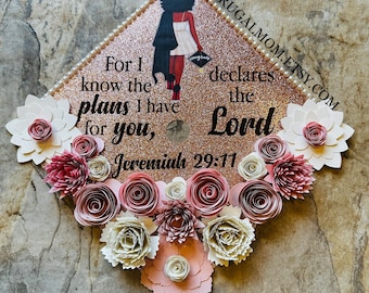 May include: A graduation cap decorated with pink and white paper flowers. The cap is rose gold glitter and features a black silhouette of a woman in a graduation gown. The text "For I know the plans I have for you, declares the Lord Jeremiah 29:11" is printed on the cap.