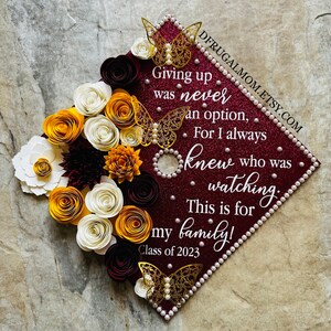 Könnte beinhalten: Eine bordeauxfarbene Abschluss-Kappe, verziert mit weißen, goldenen und kastanienbraunen Papierblumen, goldenem Glitzer und Perlen. Die Kappe trägt den Text "Giving up was never an option, For I always knew who was watching. This is for my family! Class of 2023".