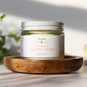 May include: A small glass jar with a silver lid, labeled "JJLautz Unscented Tallow Cream." The jar contains a creamy substance and is set on a round, wooden dish. The label indicates the jar contains 2 oz. of cream.
