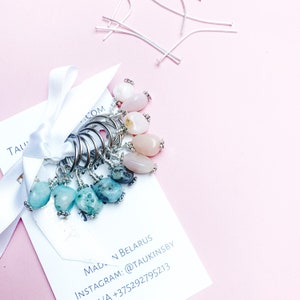 Op de afbeelding: Set van 10 zilveren ringvormige flessenstoppers met verschillende roze en blauwe edelsteenbedels. De bedels zijn met zilverdraad aan de ringen bevestigd. De stoppers worden gepresenteerd op een witte kaart met de tekst "TAU...COM" en contactgegevens.