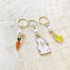 Puede incluir: Tres marcadores de punto de oro con cuentas de perla y encantos. Los encantos son una zanahoria, un conejito y un pollito.