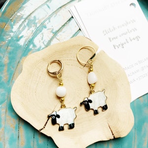 Op de afbeelding: Een paar gouden schapenoorbellen met wit en zwart email. Elke oorbel heeft een witte kraal en een gouden ring.