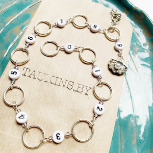 以下が含まれることがあります： 0から9までの数字が書かれた白いビーズと小さな貝殻のチャームが付いたシルバーチェーンネックレス。ネックレスは、「TAUKINS.BY」という文字が印刷された茶色の表面の上に置かれています。