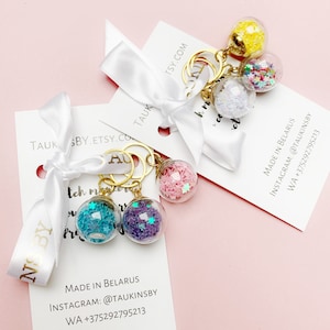 May include: A set of four gold keychains with glass charms filled with colorful glitter. Each charm has a different color glitter: pink, blue, purple, and yellow. The keychains are packaged on white cards with the text "TAUK BY.ETSY.COM" and "MADE IN BELARUS" along with contact information.