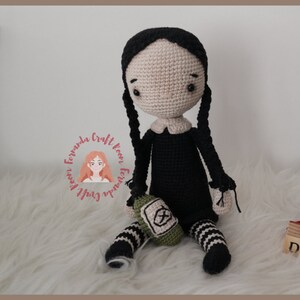 May include: A crocheted doll with black hair in pigtails, wearing a black dress with a white collar and black and white striped stockings. The doll is holding a green crocheted bag with a white symbol. The doll is sitting on a white fluffy surface.
