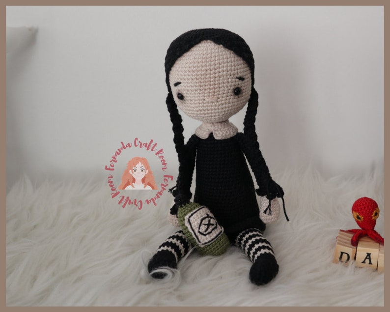 May include: A crocheted doll with black hair in pigtails, wearing a black dress with a white collar and black and white striped stockings. The doll is holding a green crocheted bag with a white symbol. The doll is sitting on a white fluffy surface.