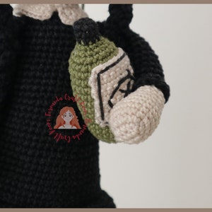 May include: Crocheted amigurumi doll wearing a black dress and holding a green crocheted bottle.
