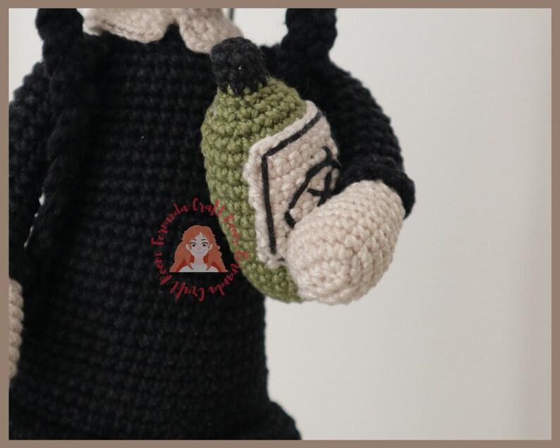 May include: Crocheted amigurumi doll wearing a black dress and holding a green crocheted bottle.