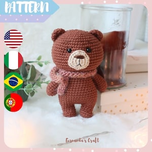 Può includere: Un orsacchiotto di peluche marrone all'uncinetto che indossa una sciarpa rosa e marrone. L'orso ha un viso beige con occhi neri e un piccolo naso nero.