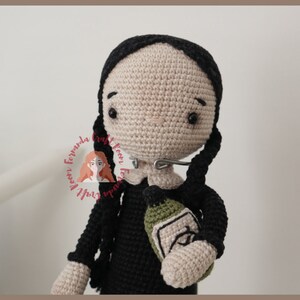 May include: Crocheted doll with black hair and a black dress holding a green bottle. The doll has a neutral expression and is wearing a silver nose ring.