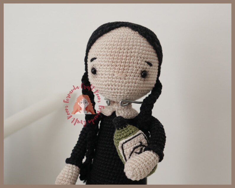 May include: Crocheted doll with black hair and a black dress holding a green bottle. The doll has a neutral expression and is wearing a silver nose ring.