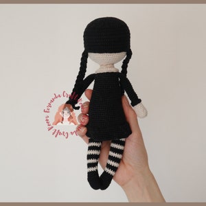 May include: Black crochet doll with white stripes on the legs and black pigtails. The doll is wearing a black dress.