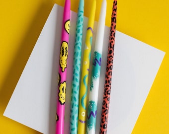 May include: Five colorful birthday candles with different patterns. The candles are decorated with geometric shapes, a smiley face, and a leopard print.
