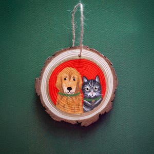 May include: A wooden slice ornament featuring a painted golden retriever and a gray tabby cat, both wearing green collars, set against a red background. The ornament is round with a natural wood grain edge and a twine loop for hanging.