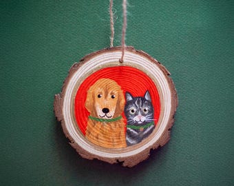May include: A wooden slice ornament featuring a painted golden retriever and a gray tabby cat, both wearing green collars, set against a red background. The ornament is round with a natural wood grain edge and a twine loop for hanging.