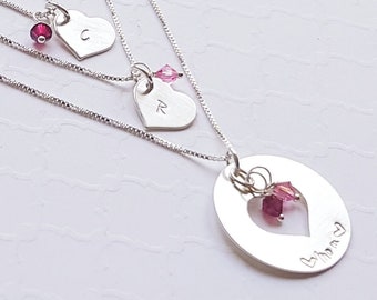 May include: Three silver necklaces with heart-shaped pendants. The top necklace has a small heart with the letter "C" on it. The middle necklace has a slightly larger heart with the letter "R" on it. The bottom necklace has a larger oval pendant with a heart cut out of the centre and the word "Mommy" engraved on it. Each necklace has a small pink crystal charm.