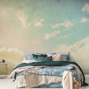 May include: Bedroom with a sky-themed mural. The wall features a gradient of colors, transitioning from yellow to blue, with fluffy white clouds. A bed with teal and beige bedding is in front of the mural. A gold side table and a basket are in the foreground.