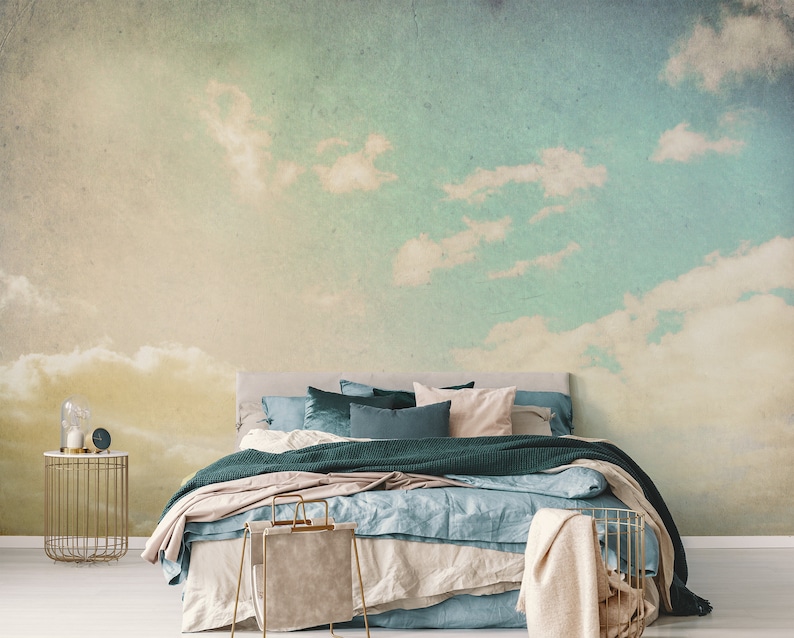 May include: Bedroom with a sky-themed mural. The wall features a gradient of colors, transitioning from yellow to blue, with fluffy white clouds. A bed with teal and beige bedding is in front of the mural. A gold side table and a basket are in the foreground.