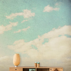 May include: A light wood cabinet with two drawers, open shelving, and a decorative geometric design. The cabinet is set against a backdrop of a sky with clouds. A woven lamp, two small blue boxes, and two wooden cups are on the cabinet.