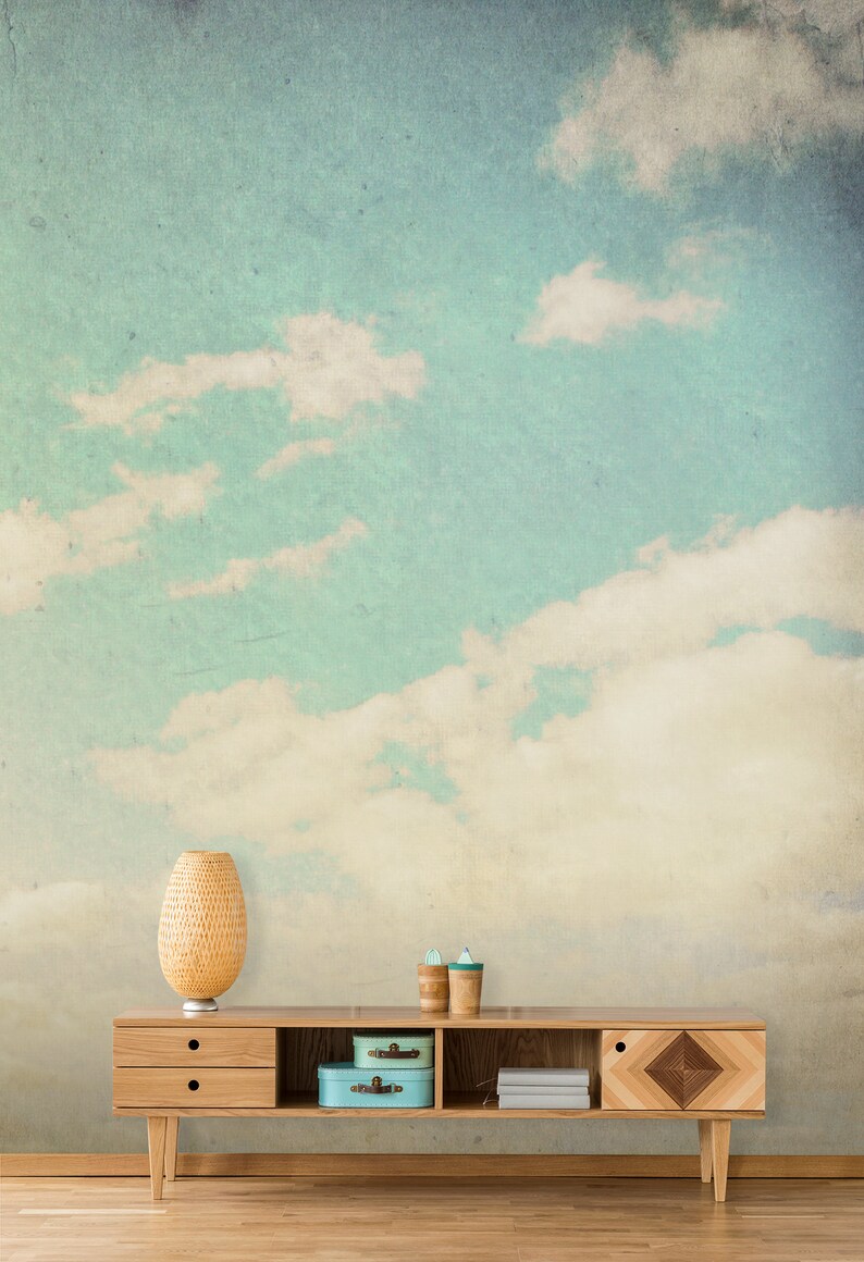 May include: A light wood cabinet with two drawers, open shelving, and a decorative geometric design. The cabinet is set against a backdrop of a sky with clouds. A woven lamp, two small blue boxes, and two wooden cups are on the cabinet.